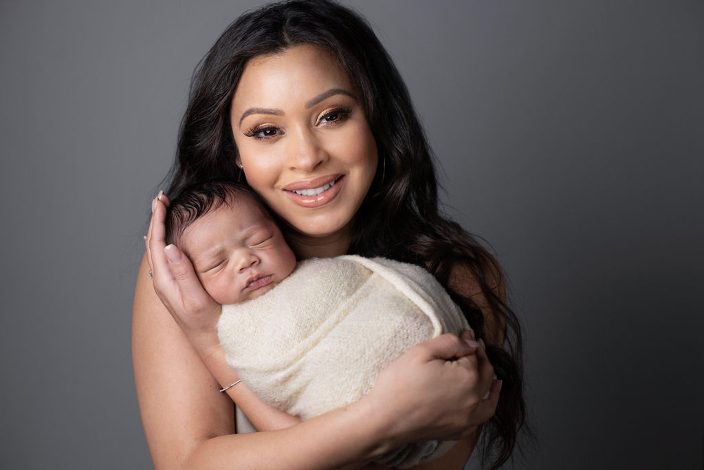 A mother smiling at the camera while holding her swaddled newborn close, against a grey backdrop.