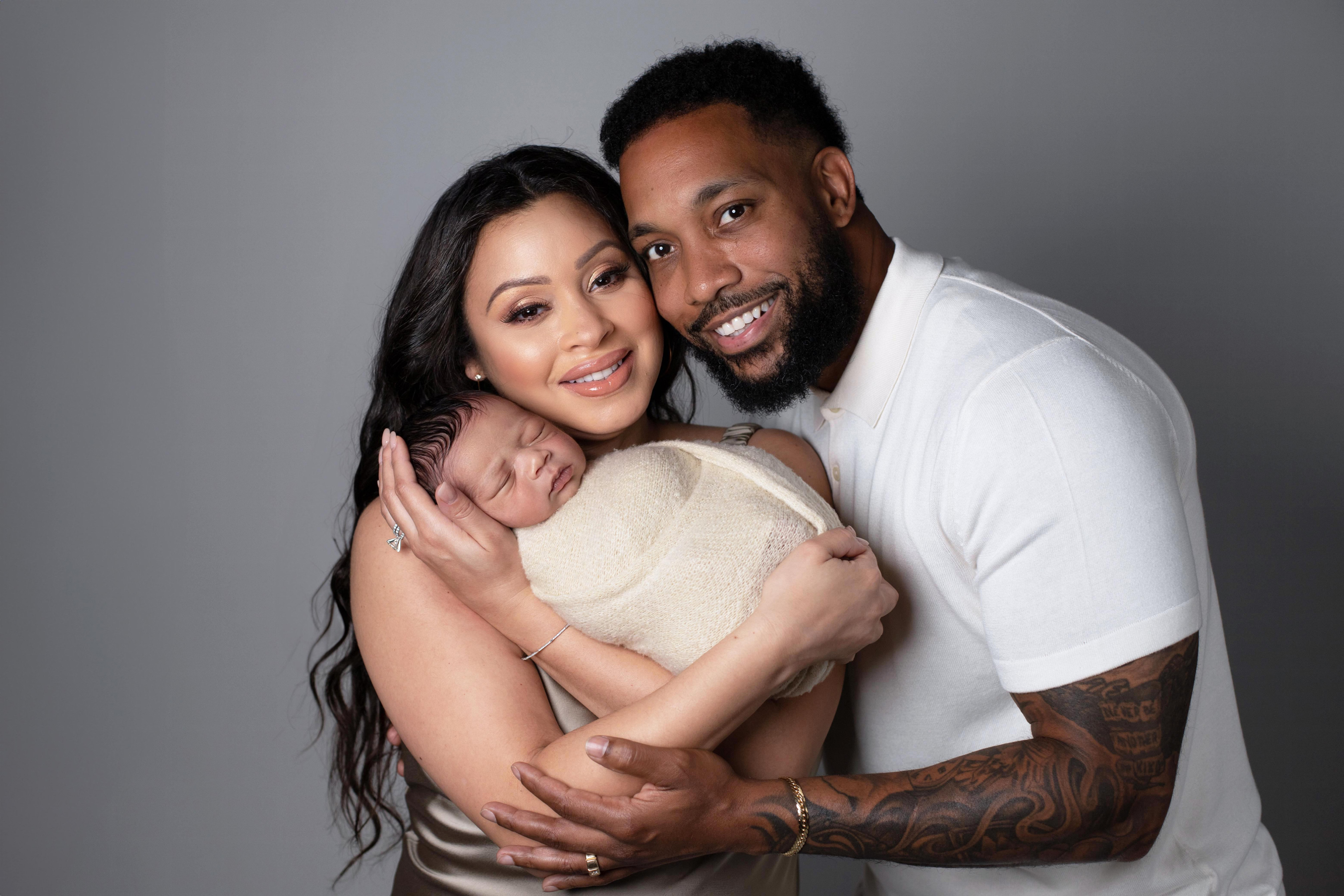 A mother and father smiling at the camera while holding their swaddled newborn between them, against a soft grey backdrop.