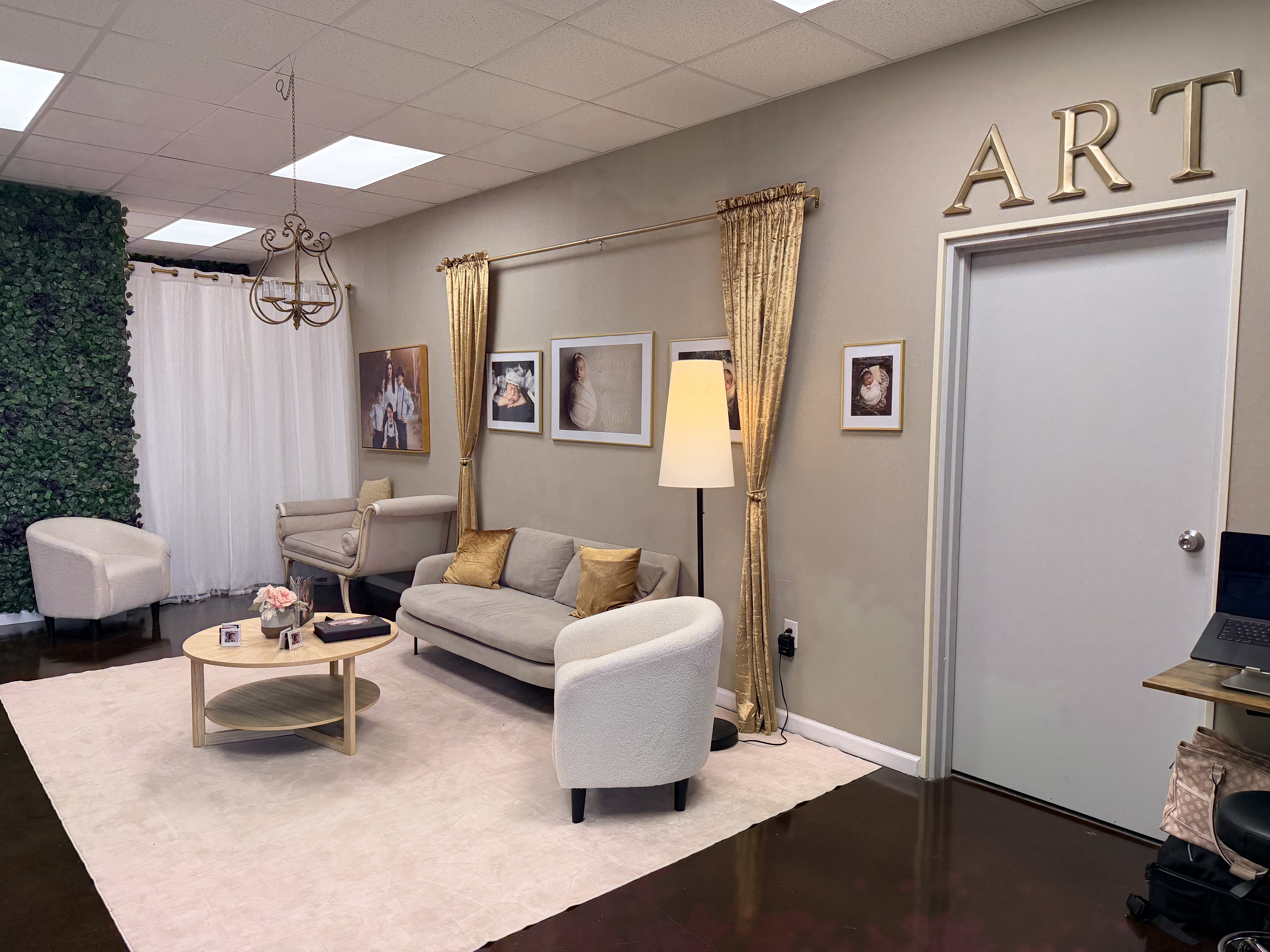 The Studio Realms reception lounge: a sage couch with gold accent pillow, gold-framed newborn portrait prints on the walls, gold curtains and chandelier, and gold ART lettering on the wall above the door.