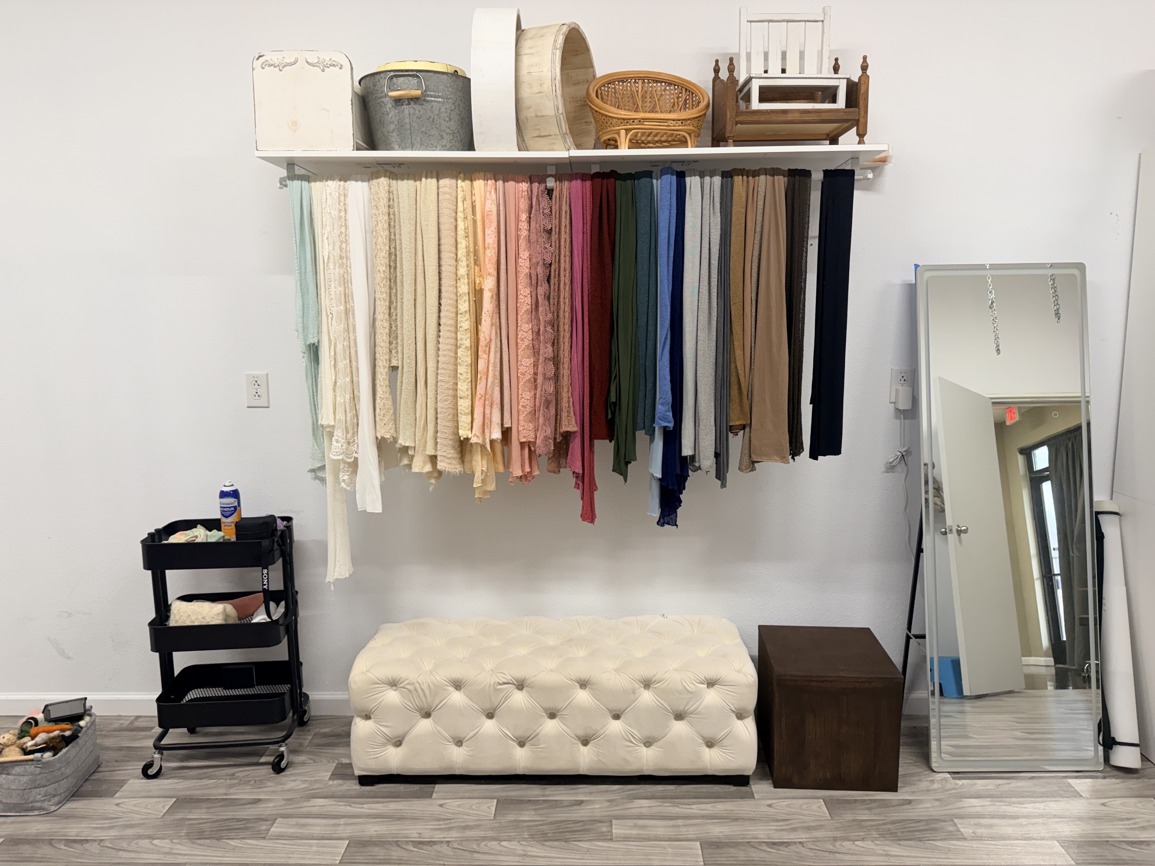 A long wardrobe rack in the Studio Realms studio with hanging fabrics in cream, blush, sage, dusty rose, navy and taupe, with woven prop baskets above and a tufted cream ottoman bench below.
