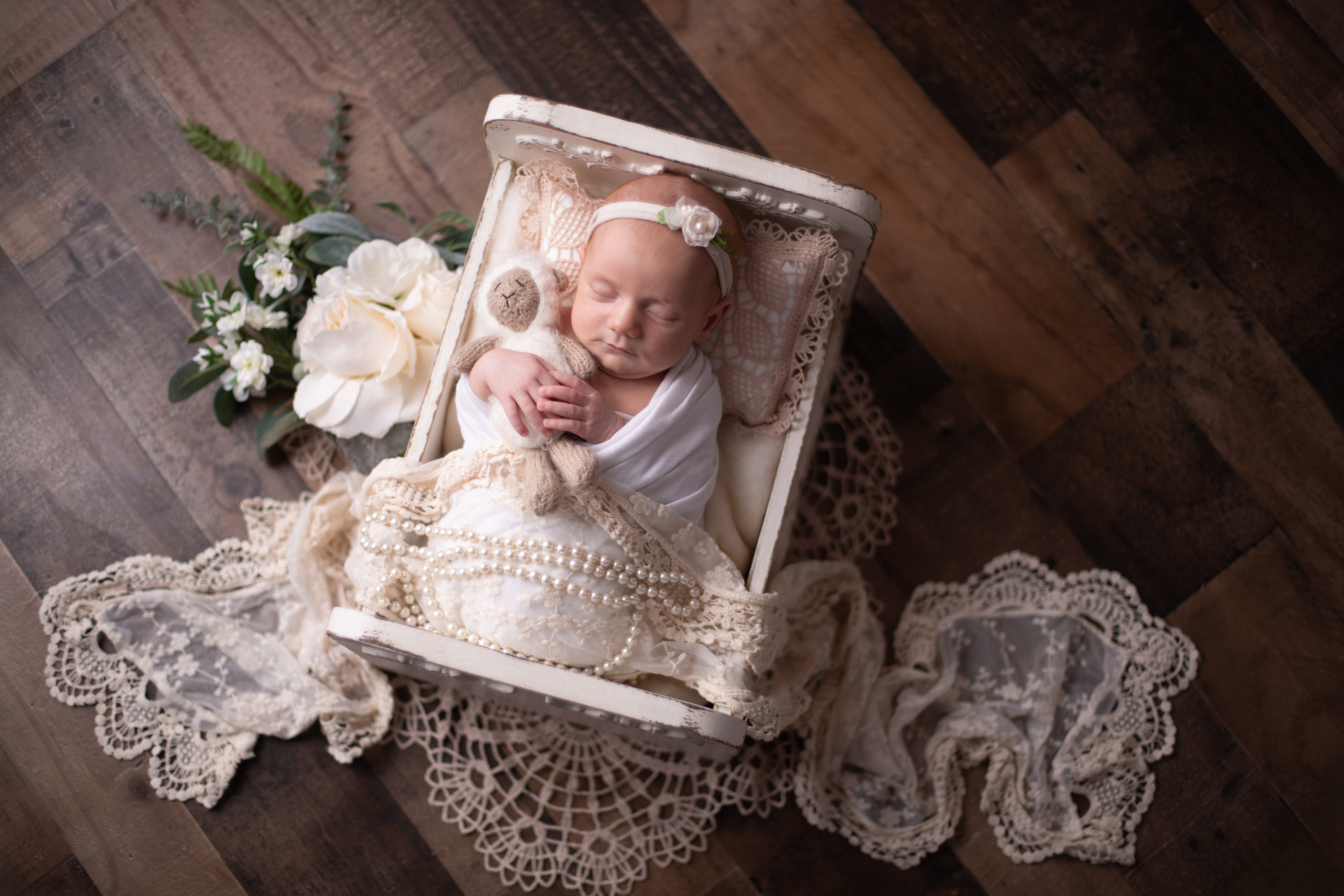 A top-down view of a swaddled newborn in cream lace with a small bow headband, posed in a small wooden bed surrounded by white florals and lace, on a warm wood plank floor.