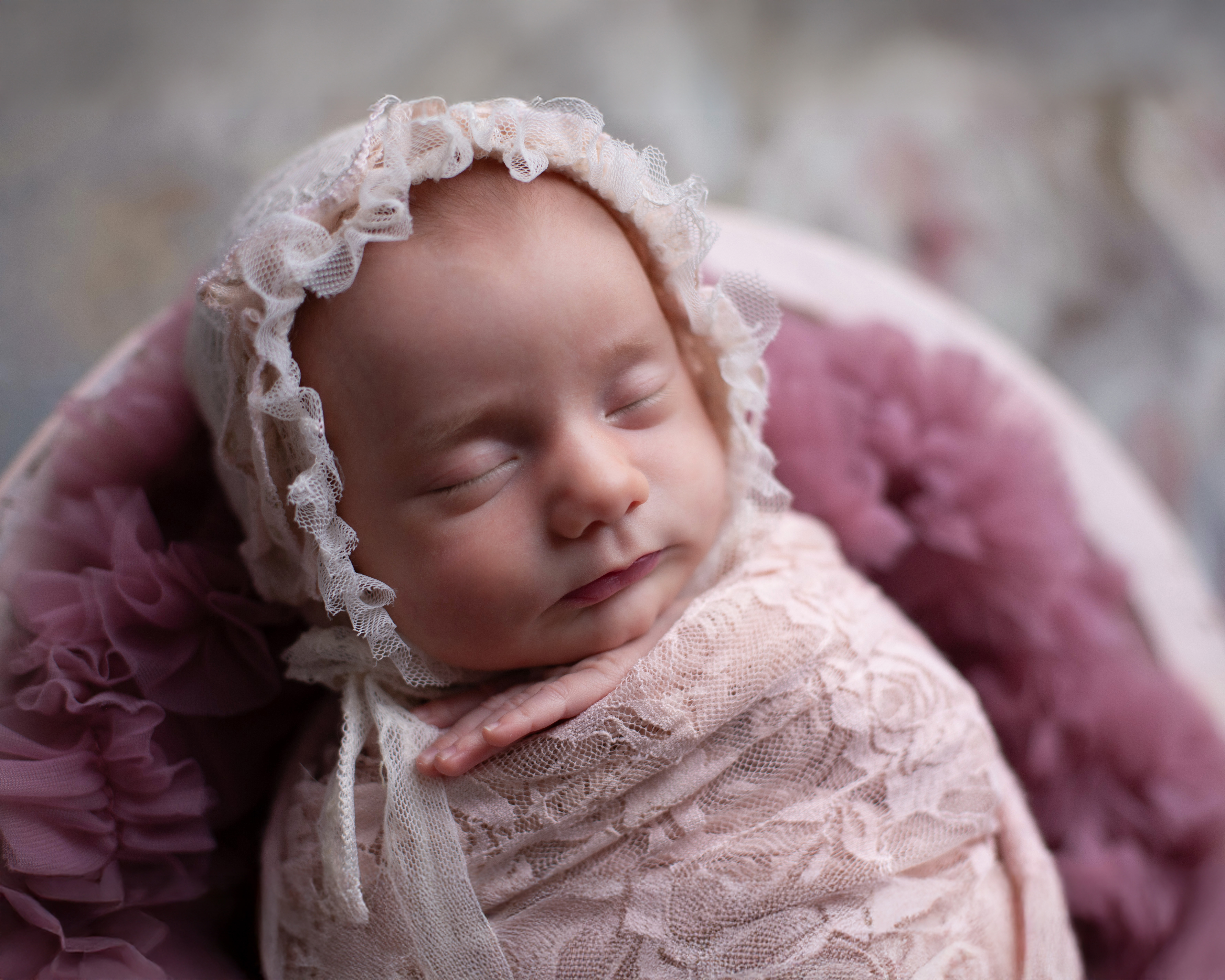 A sleeping newborn in a delicate cream lace bonnet, swaddled in cream lace, surrounded by a dusty mauve fluffy blanket against a soft pink and grey painted backdrop.