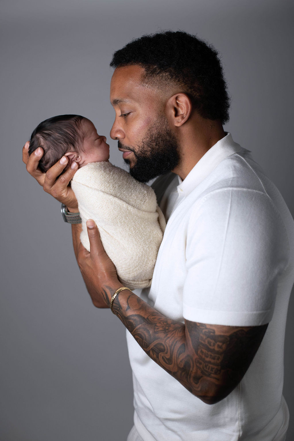 A father in a white polo shirt seen in profile, kissing the top of his swaddled newborn's head, holding the baby close to his face, against a grey backdrop.