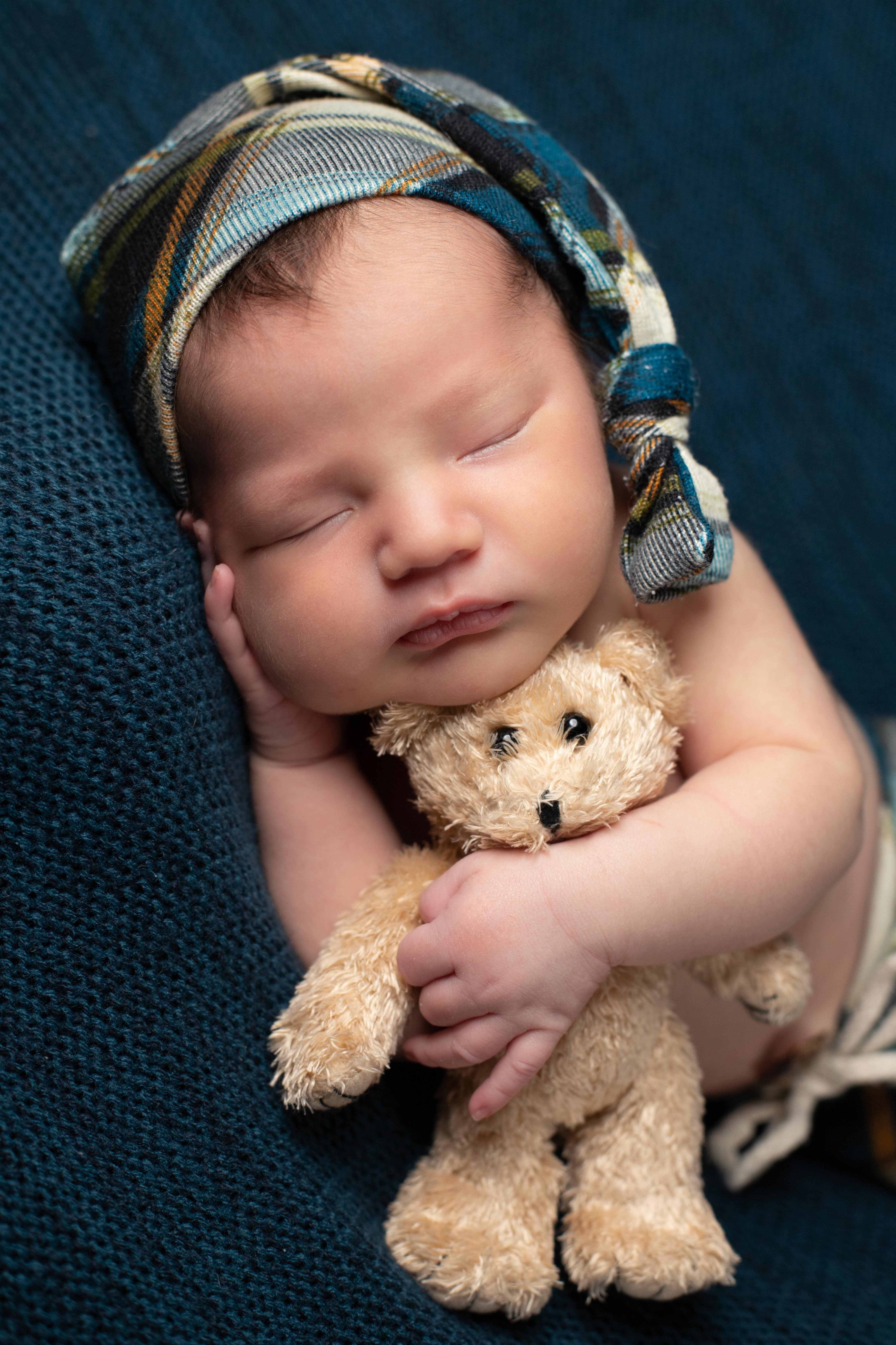 A sleeping newborn in a plaid blue and tan bonnet, holding a small teddy bear on a navy textured blanket.
