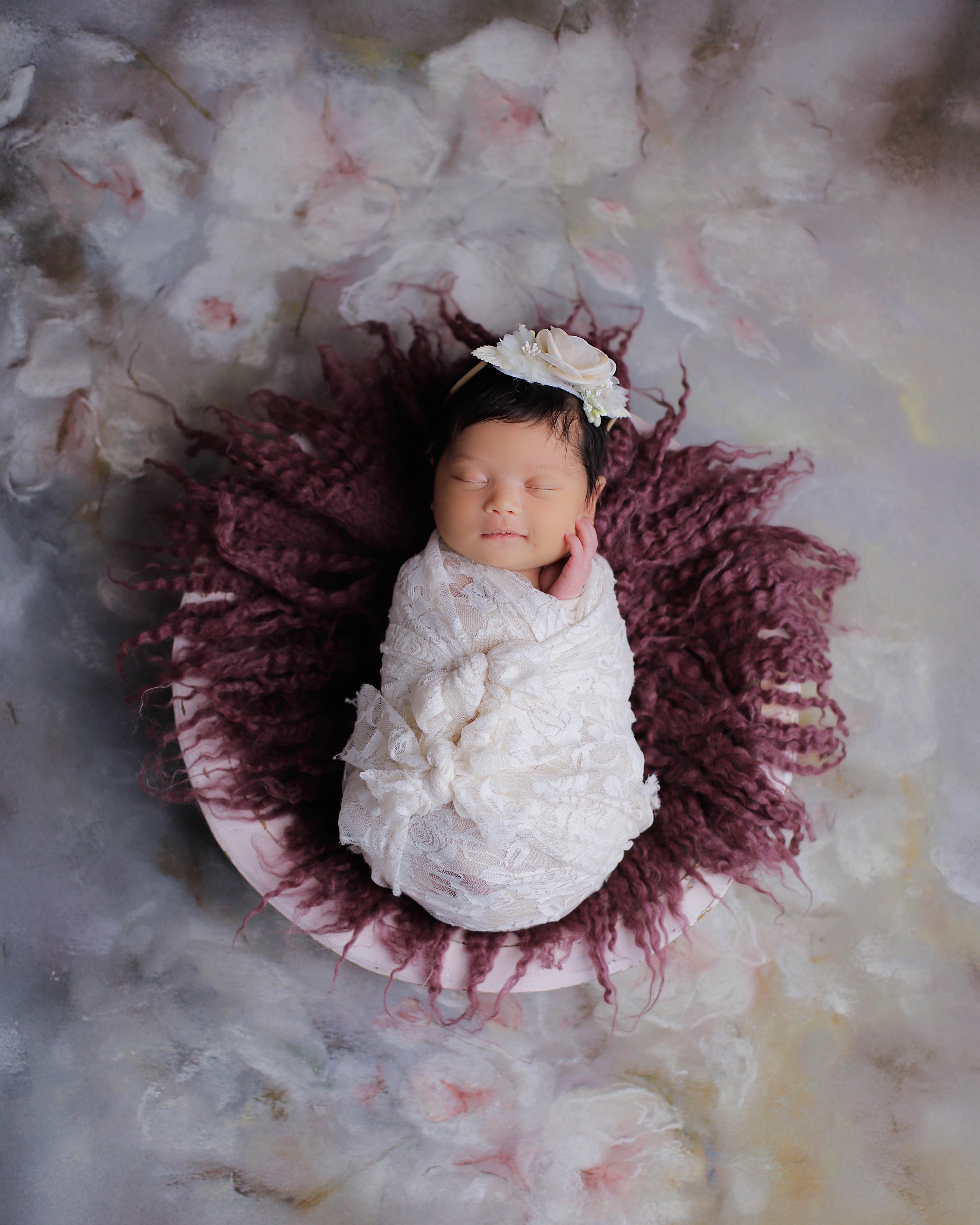 A top-down view of a swaddled newborn in white lace, posed in a small bowl lined with maroon yarn, surrounded by white cotton bolls on a soft pink-and-cream painted backdrop.