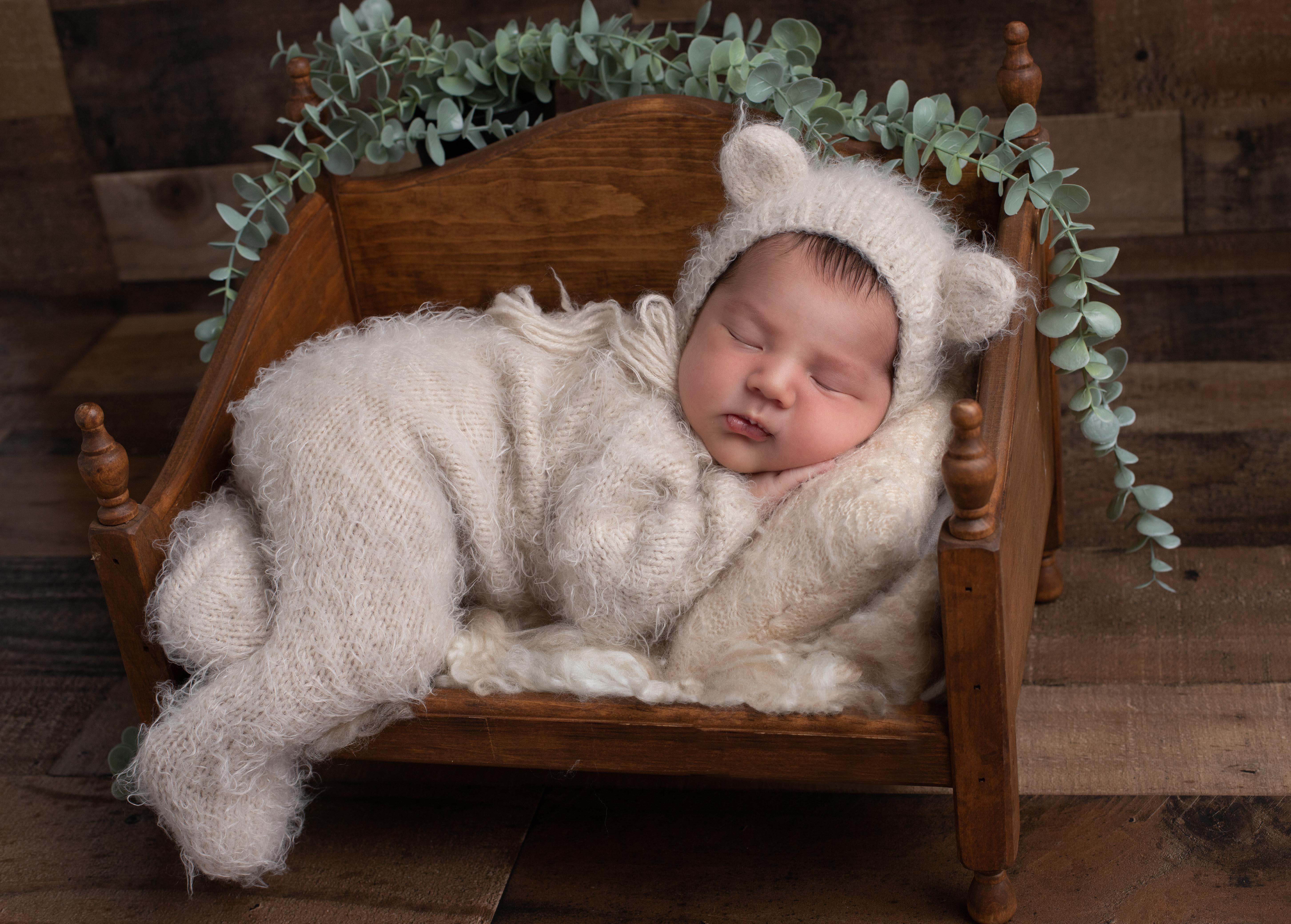 A sleeping newborn in a cream knit bear bonnet and matching cream knit outfit, posed in a small wooden bed prop with eucalyptus greenery, against a wood plank backdrop.