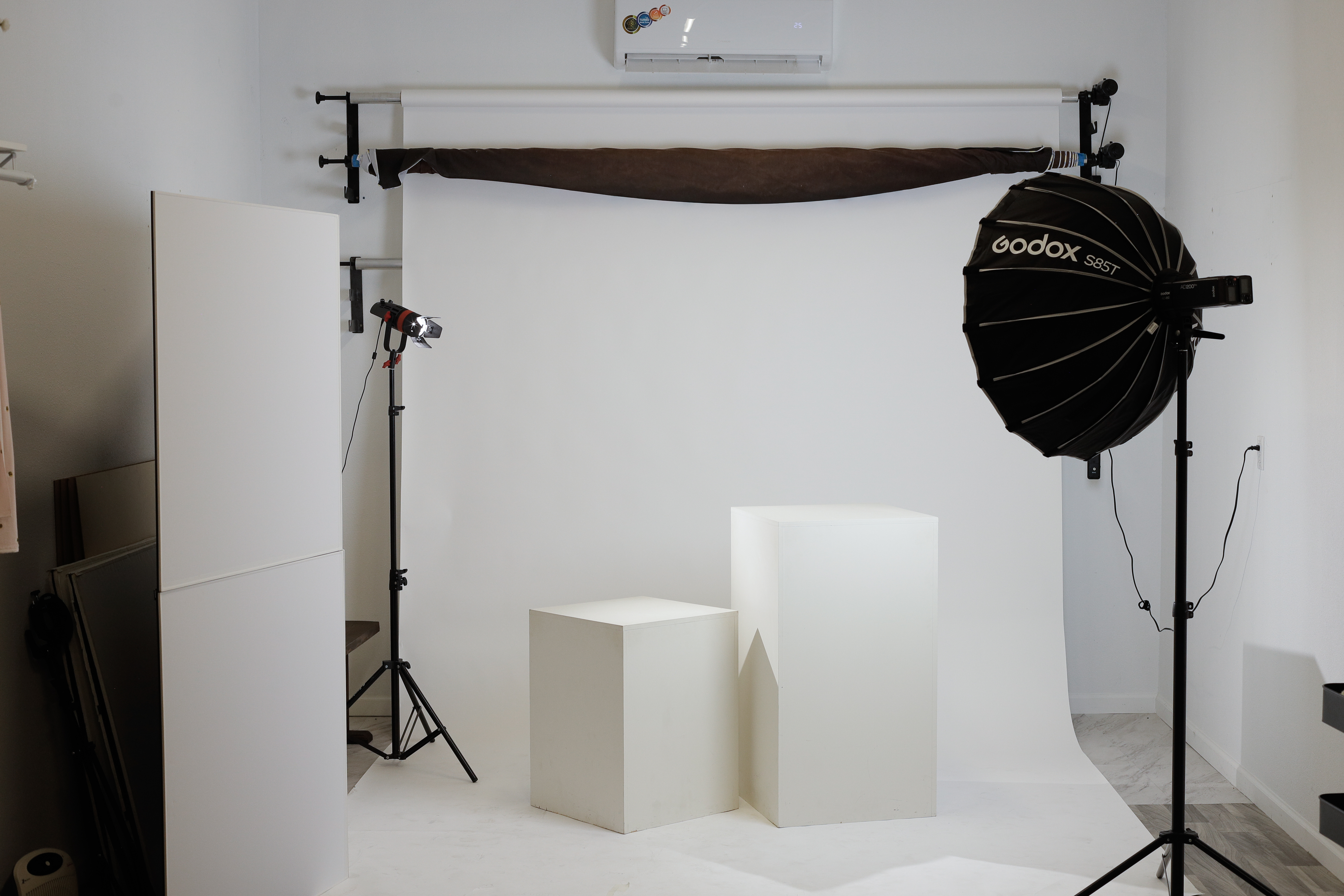 The Studio Realms shooting space: a white seamless backdrop with a brown muslin draped across the top, two white posing platforms, a Godox S85T softbox on a stand to the right, and a continuous light to the left.