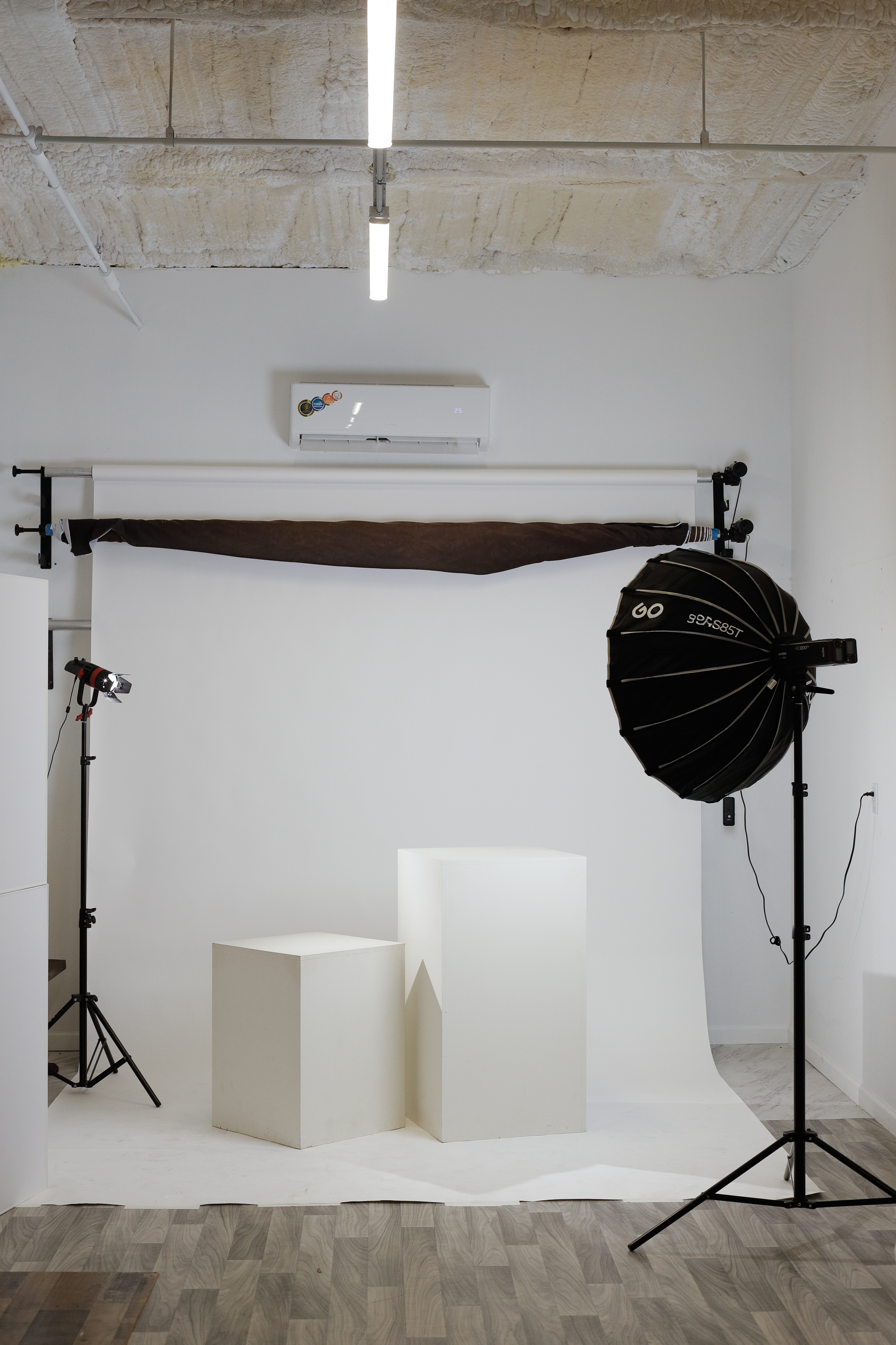 The Studio Realms shooting setup: a white seamless backdrop with a brown muslin pulled across the top, two posing platforms in front, a Godox S85T softbox on a stand to the right, a continuous light to the left, and exposed concrete ceiling above.