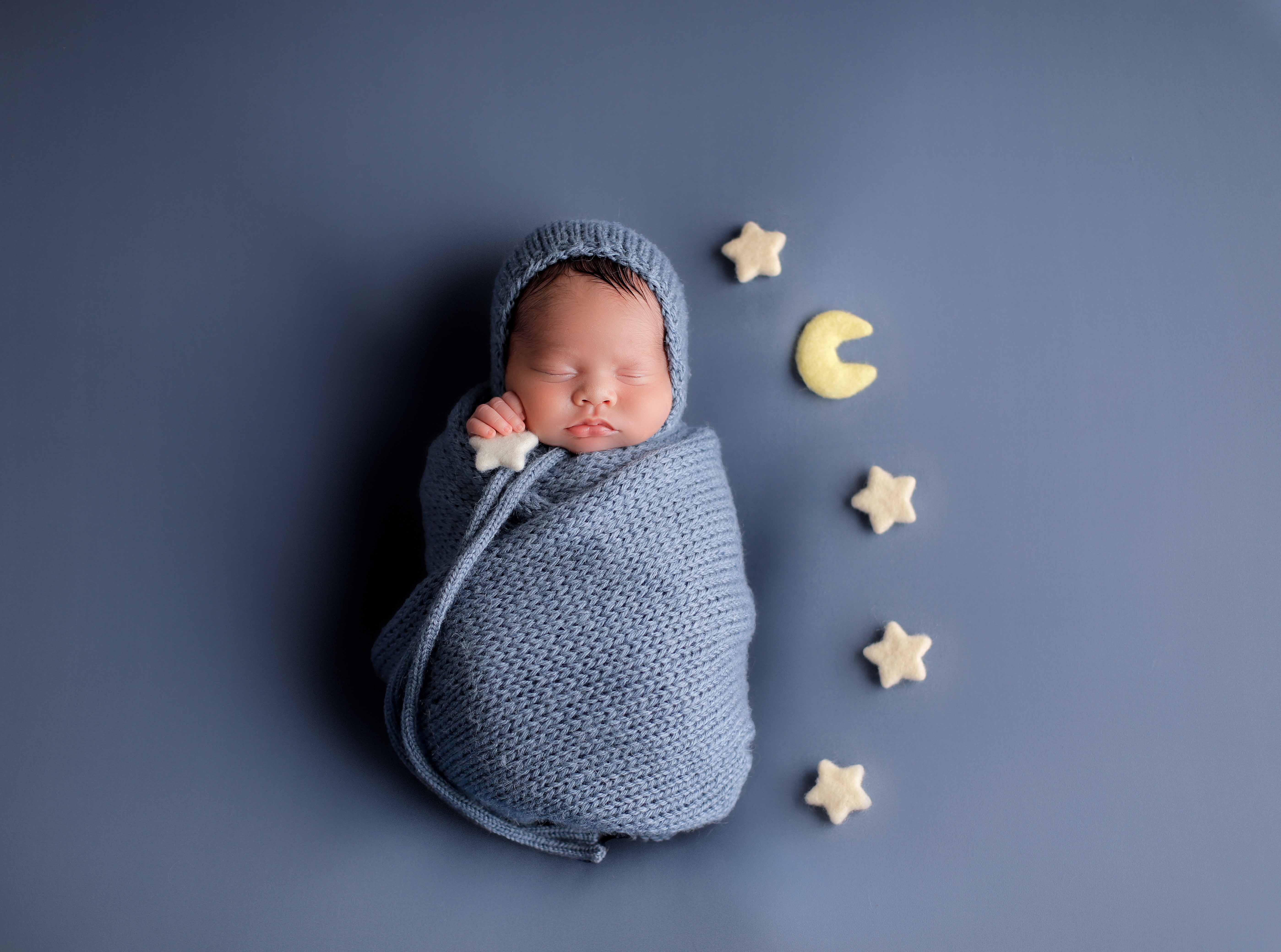 A swaddled newborn in a soft blue knit wrap and matching bonnet, sleeping on a dusty blue backdrop with felt moon and stars props arranged beside the baby.